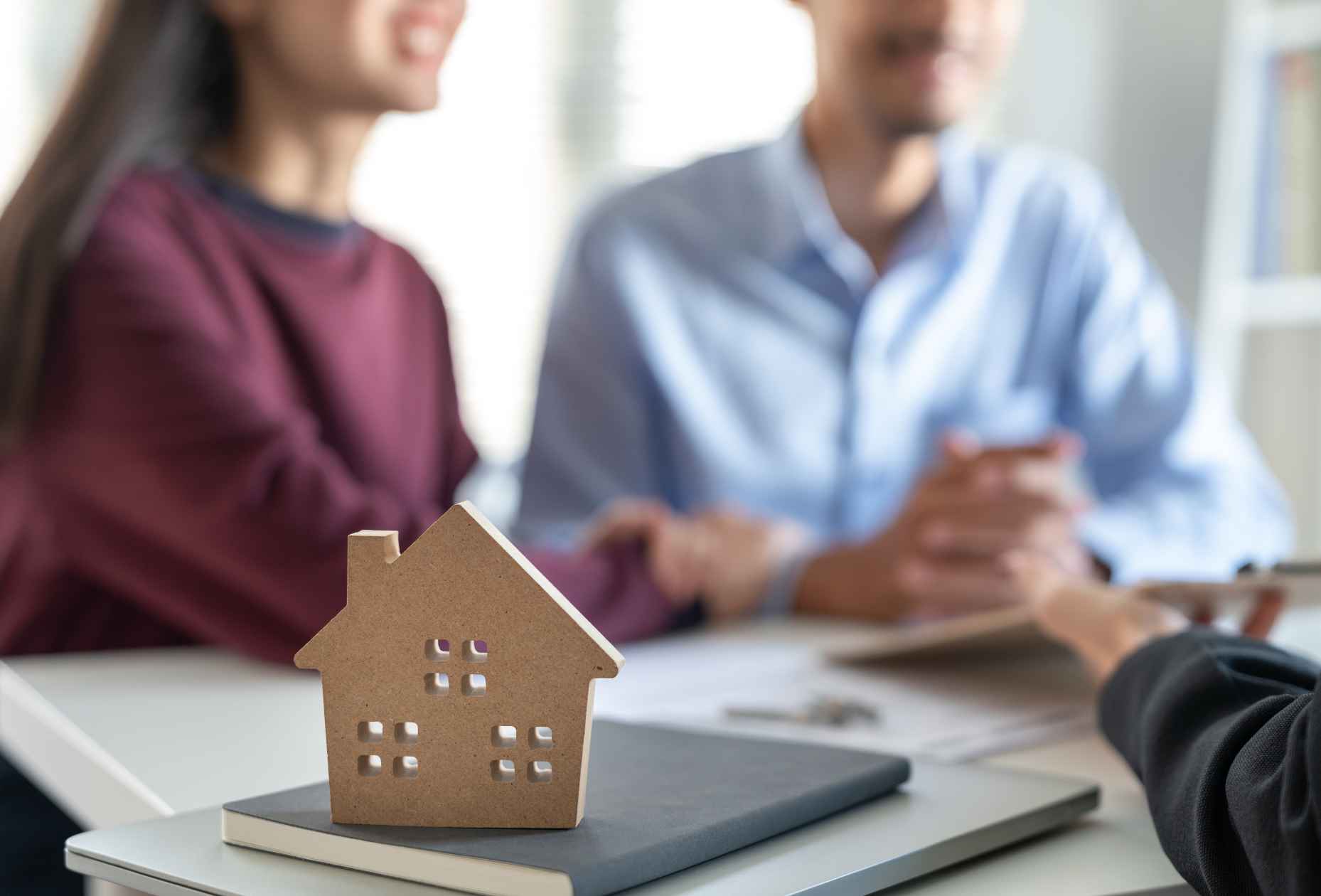 A young newly married couple sits across from a real estate agent in an office, discussing property options, joint homeownership benefits, and home loan eligibility.